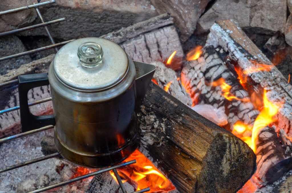 A vintage percolator sits over a campfire, its contents nearly ready as the coals glow fiercely beneath it. This image captures the quintessential essence of outdoor brewing, a tradition that harkens back to bygone days. The scene symbolizes not just the act of making coffee but a cherished ritual that enriches the experience of each cup with history, aroma, and the rich flavor of coffee made with fire, patience, and a touch of nostalgia.