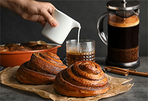 A hand pouring cream into a glass of coffee, with a French press and freshly baked cinnamon rolls in the background, embodying the delightful experience of enjoying a rich and complex cup of French press coffee paired with sweet pastries.