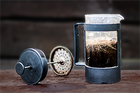 The unassembled parts of a French press laid out on a wooden surface, showing the carafe and the plunger with its metal filter, highlighting the simplicity of the design and the eco-friendly aspect of its construction that minimizes waste.