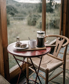 A serene morning coffee scene featuring a French press, cup, and pastries on a rustic bistro table with a view of the countryside through a glass door, exemplifying the simplicity and charm of brewing with a French press for beginners.
