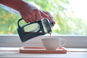 A hand pouring steaming coffee from a French press into a cup, set upon a light wooden tray by a bright window, encapsulating the step-by-step journey of brewing the perfect cup of coffee with a French press, from grinding and steeping to the enjoyment of its rich, full flavor.