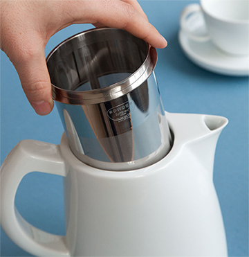 : A hand placing the stainless steel filter basket into the Sowden SoftBrew Coffee Maker, a key step in the immersion brewing process.