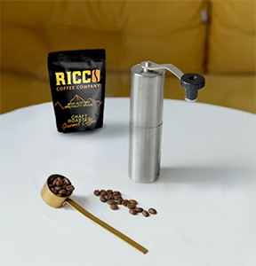 A pack of Ricco Coffee Company beans alongside a manual coffee grinder and a wooden scoop with beans, ready for grinding to the ideal coarse size for a French press, on a white table against a soft yellow background.