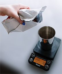 Pouring Ricco Coffee Company beans into a grinder on a digital scale, capturing the precision needed for a consistent French press coffee ratio and highlighting the importance of measuring tools in the brewing process.