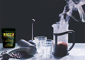 Steam rises from a French press beside a bag of Ricco Coffee Company beans and a stainless steel burr grinder, capturing the ideal preparation for a richly flavored brew.