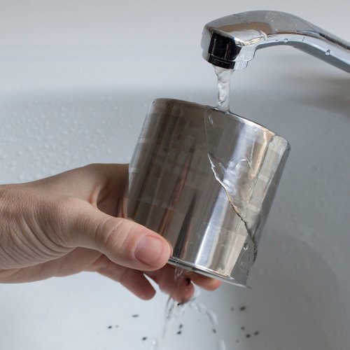 A hand washing a stainless steel filter of a Sowden coffee maker under running tap water, highlighting the simplicity of the cleaning process.