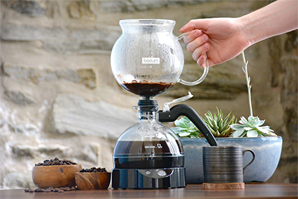 A hand removing the top chamber from a Bodum siphon vacuum coffee maker after brewing, with coffee beans and a succulent plant in the background, encapsulating the full siphon coffee experience.
