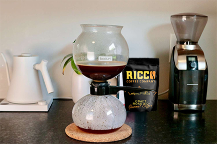 A Bodum siphon vacuum coffee maker in the midst of brewing, accompanied by a bag of Ricco Coffee Company beans and a coffee grinder, representing the importance of using freshly ground coffee and maintaining equipment for the best coffee experience.