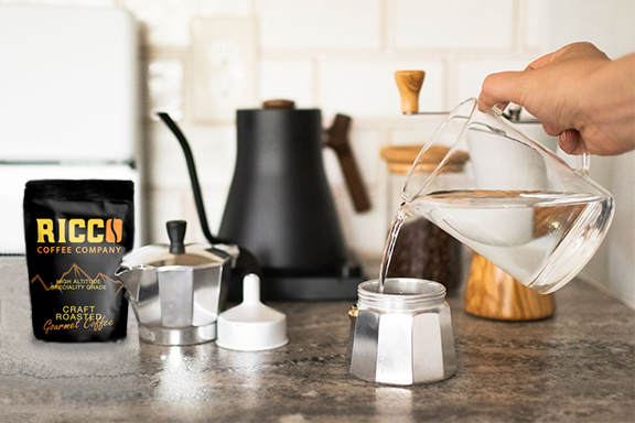 The beginning of the brewing process with a Moka Pot, a hand pouring water, alongside a bag of Ricco Coffee Company's artisan craft - roasted beans on a kitchen countertop.